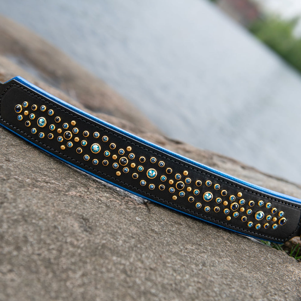 A black leather dog collar with blue trim and blue crystals in brass hardware, photographed resting on a large stone with water and trees blurred in the background.