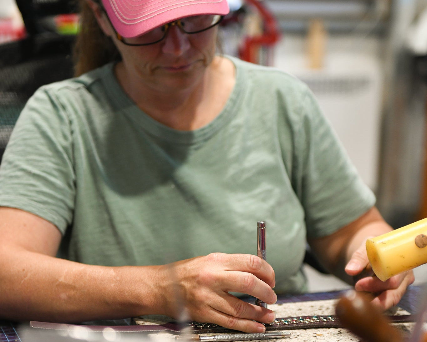 A woman wearing a pink cap and glasses sits at a workbench, holding a hand tool, crafting a leather dog collar.  The background shows a workshop environment.