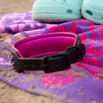 Pink and purple towel with a black dog collar and light blue sandals on sand
