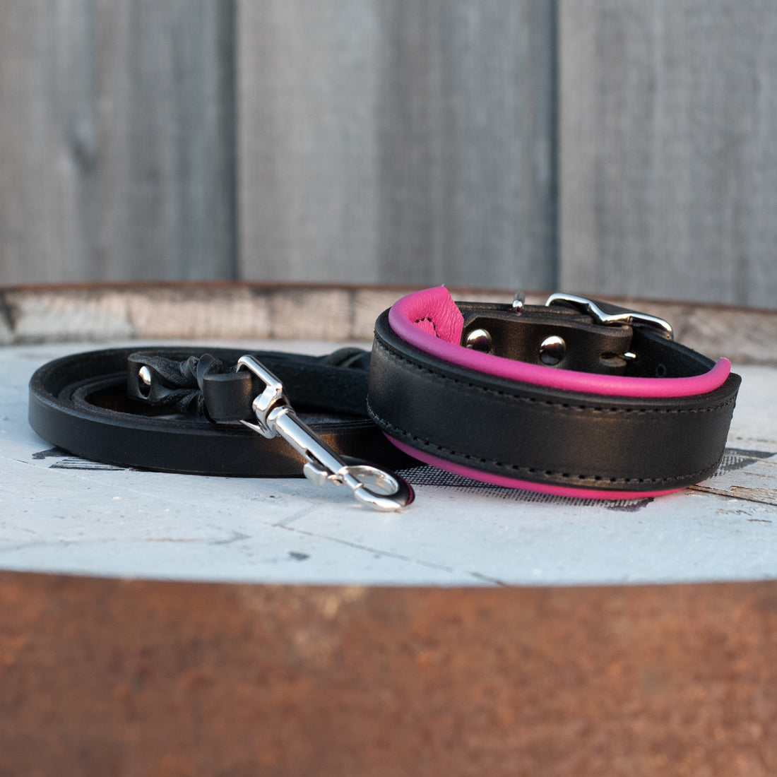A black leather dog collar with a bright pink inner lining and a matching black leash with silver metal hardware, displayed on a rustic wooden surface.