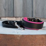 A black leather dog collar with a bright pink inner lining and a matching black leash with silver metal hardware, displayed on a rustic wooden surface.