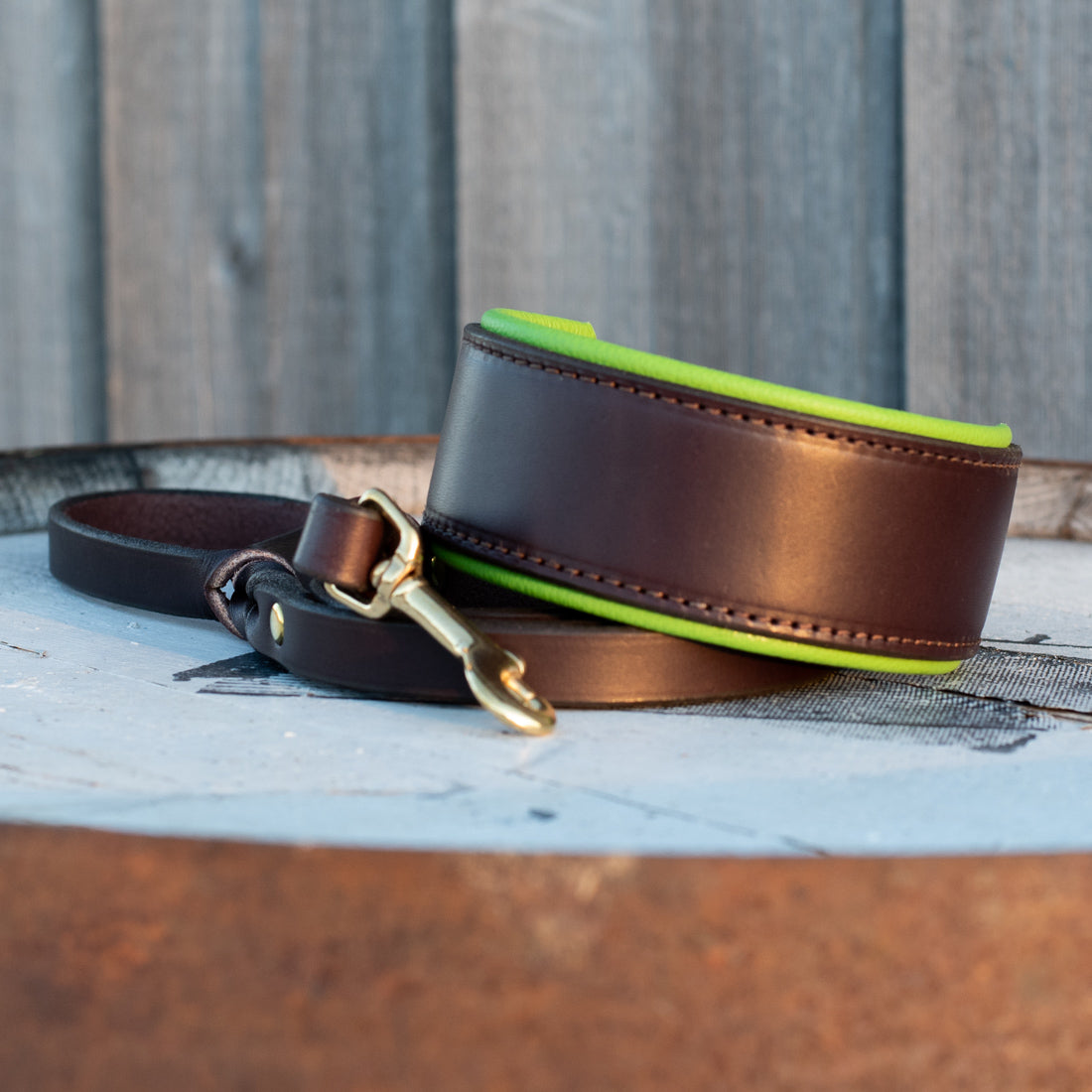 A dark brown leather dog collar with a bright lime green inner lining and a brass-tone snap hook, resting on a weathered wooden surface with a gray wood plank background.