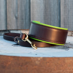 A dark brown leather dog collar with a bright lime green inner lining and a brass-tone snap hook, resting on a weathered wooden surface with a gray wood plank background.