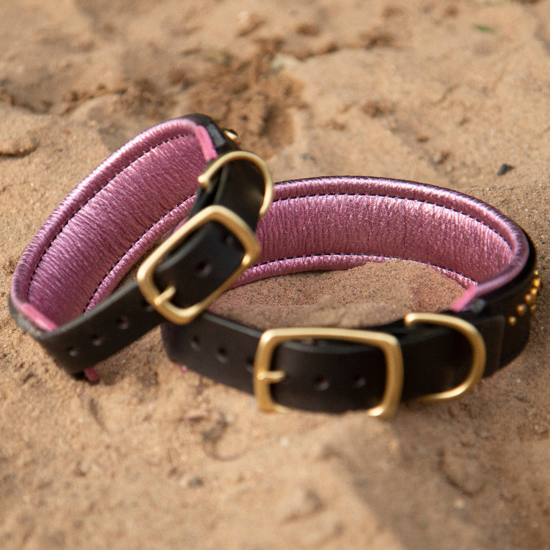 Two Black leather dog collars with metallic pink padding on a sandy beach