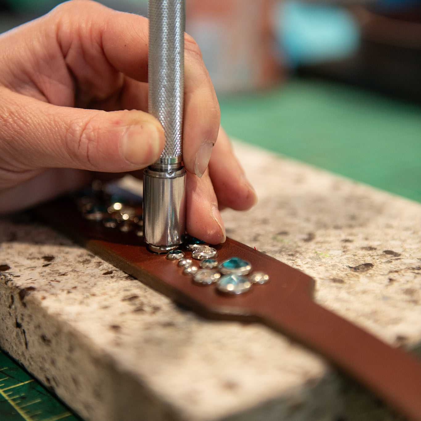 Hand using a tool on a leather strap with decorative elements on a stone surface.