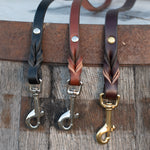 Three leather leashes in black, medium brown, and chocolate brown hang from a rustic oak barrel, each with a silver or brass snap.